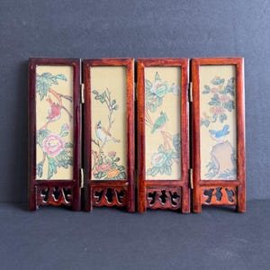 May include: A small, folding screen with four panels. Each panel displays a painted bird scene with flowers and foliage on a cream background. The frames are a dark reddish-brown colour, and the screen has decorative feet.
