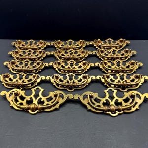 May include: A collection of twelve brass-toned metal drawer pulls arranged in four rows of three. Each pull features an ornate, openwork backplate with a curved, swinging handle attached to two small circular mounting points on the decorative frame.