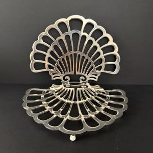 Puede incluir: Un salvamanteles de metal calado de color plateado, con diseño de abanico o concha. El salvamanteles tiene un borde festoneado y un patrón calado decorativo. Tiene un pequeño pie blanco. El fondo es negro.