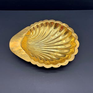 May include: A decorative, golden-colored seashell-shaped dish. The dish has a scalloped edge and a textured, ridged interior. The overall design is reminiscent of a natural seashell, with a polished, reflective surface.