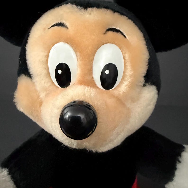 May include: Close-up of a plush Mickey Mouse toy. The toy features a black and white color scheme with a large black nose, white eyes with black pupils, and black ears. The face is tan and the body is black.