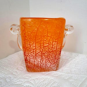 Puede incluir: Jarrón de cristal naranja con un patrón texturizado y agrietado. El jarrón tiene forma cuadrada con dos asas de cristal transparente. El color naranja es vibrante y el diseño le da un aspecto artístico único. Una pieza decorativa para la decoración del hogar.