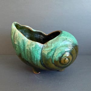 May include: A ceramic planter shaped like a seashell, with a glossy green and brown glaze. The planter has a scalloped rim and rests on three small feet. The spiral design is prominent.