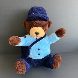 May include: A plush teddy bear wearing a blue uniform and hat. The bear has brown fur on its head, arms, and legs, with a light blue shirt and dark blue pants. It has a light tan muzzle and a black nose. The bear is sitting with its arms outstretched.