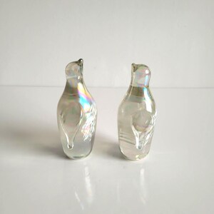 May include: Two iridescent glass decorative bottles with a pearlescent finish. The bottles have a curved shape with a small opening at the top and a teardrop-shaped design on the front. The glass reflects a rainbow of colors.