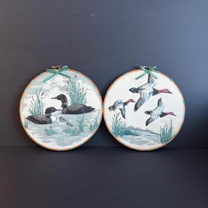 Peut inclure: Deux décorations murales circulaires avec des impressions sur tissu représentant des oiseaux aquatiques. La pièce de gauche montre deux huards adultes et deux poussins dans un étang. La pièce de droite montre trois canards en vol. Bords orange et nœuds verts.