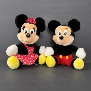 May include: Two plush Disney character dolls, Mickey and Minnie Mouse, sit side-by-side. Minnie wears a red polka-dot dress and bow. Mickey wears red shorts. Both have black fur, white gloves, and yellow feet.