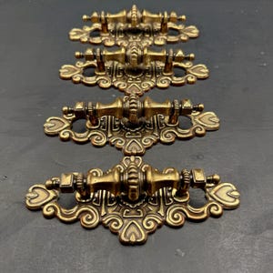 May include: Four antique brass drawer pulls arranged in a vertical column. Each pull features a central horizontal handle with a ribbed design, mounted on an ornate, scroll-patterned backplate with heart-shaped cutouts at the ends.