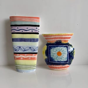 May include: Two colorful ceramic vases. The taller vase is rectangular with horizontal stripes in orange, black, purple, yellow, and blue, with decorative patterns. The shorter vase is round with a blue square design and a flower in the center, with orange and yellow stripes.
