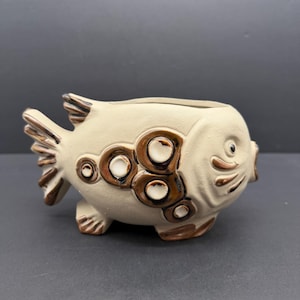 May include: A ceramic fish-shaped planter in a cream color with brown accents. The fish has a wide open mouth and a large opening on top for planting. The fish has brown circles on its side and brown fins. The planter is a decorative item.