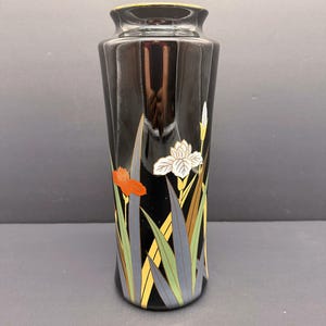 Puede incluir: Un jarrón negro con borde dorado y diseño floral. El jarrón presenta un diseño de iris estilizado con detalles en naranja, blanco y verde. El jarrón mide aproximadamente 20 cm de alto.