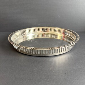 May include: A round, silver-colored serving tray with a pierced gallery rim. The tray has a reflective surface and a decorative edge. The tray is likely made of metal and is suitable for serving food or displaying items.