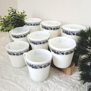 May include: Eight white ceramic coffee cups with a black floral border. Each cup has a small handle. The cups are arranged on a white patterned surface. The cups are likely for serving tea or coffee.