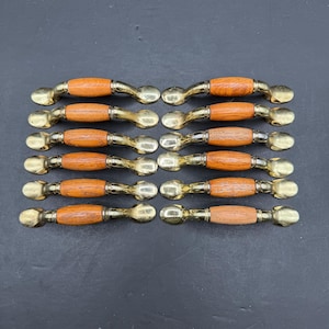 May include: A set of twelve cabinet pulls arranged in two columns. Each pull features a central cylindrical wooden handle with a visible grain, flanked by polished brass metal ends that curve outward at the mounting points for attachment.