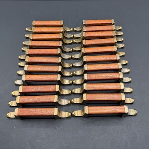 May include: A collection of eighteen cabinet pulls arranged in two columns. Each pull features a rectangular wooden centre with a visible grain pattern, flanked by two metallic brass-toned mounting brackets on either side of the handle.