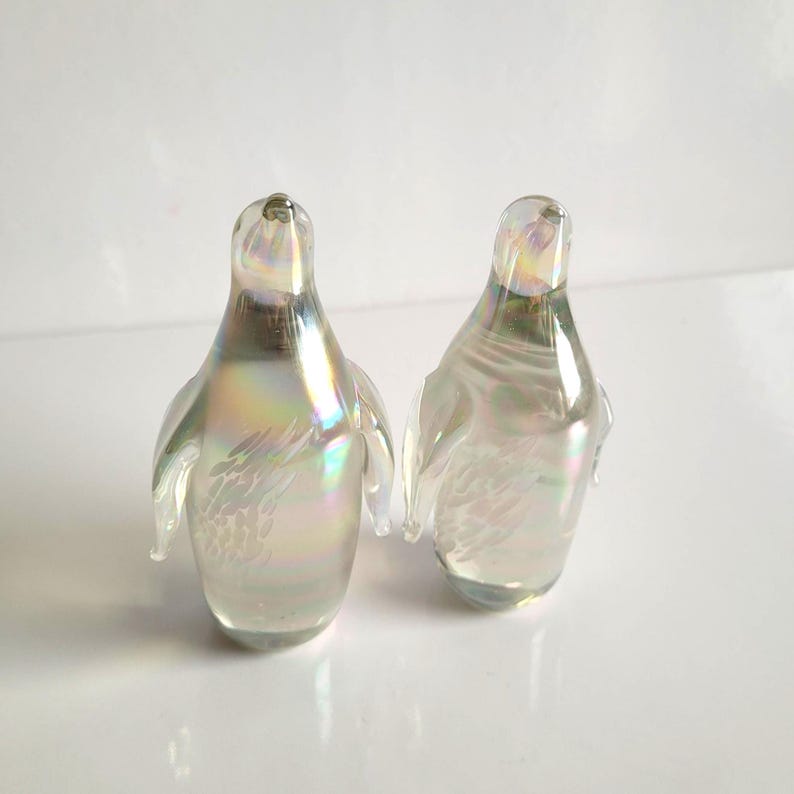 May include: Two iridescent glass penguin figurines. The penguins have a pearlescent finish with hints of yellow, green, and blue. The wings are slightly extended. The figurines are displayed on a white surface.