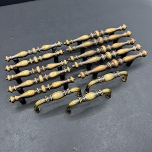 May include: A collection of ten metal drawer pulls featuring an antique brass finish. The hardware consists of eight long, slender pulls with a central spindle design and two shorter, curved pulls, all displaying a consistent metallic gold tone.