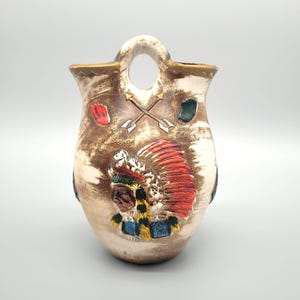 May include: A ceramic vase with a handle and a decorative Native American chief design. The vase features a brown and cream colour scheme with red, green, and blue accents. The vase has a unique shape and is suitable for home decor.