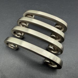 May include: Four brushed nickel cabinet pulls arranged in a parallel row. Each handle features a smooth, arched center bar that transitions into a decorative, curled scroll design at both ends, providing a consistent metallic finish throughout.