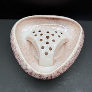 Może przedstawiać: Trójkątny ceramiczny floret z kremowym szkliwem i czerwonobrązowym wzorem spękań. Środek posiada uniesioną, perforowaną wkładkę z wieloma okrągłymi otworami przeznaczonymi do układania łodyg wewnątrz podstawy w kształcie misy.