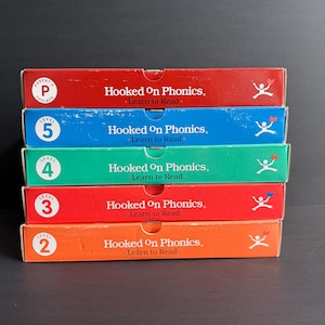 May include: A stack of five rectangular Hooked On Phonics Learn to Read boxes in red, blue, green, red, and orange. Each box features the text Hooked On Phonics Learn to Read, a level number from 2 to 5 or Parent's Toolbox, and a white stick figure icon.