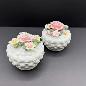 May include: Two white ceramic trinket boxes with a textured, dotted surface. Each box is topped with a cluster of colorful, detailed ceramic flowers in shades of pink, yellow, and white. The boxes are set against a dark background.