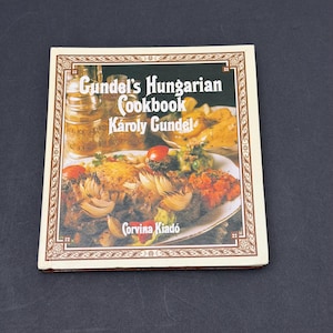 May include: A cookbook titled "Gundel's Hungarian Cookbook" by Károly Gundel. The cover features a photograph of various Hungarian dishes, including roasted vegetables and meat. The book has a cream-colored border with decorative patterns and the publisher's name, Corvina Kiadó.