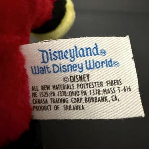 May include: A close-up of a white tag with blue text that reads "Disneyland Walt Disney World" and "&copy; DISNEY". The tag also includes text about materials and the product's origin in Sri Lanka.