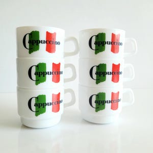 May include: Six white ceramic cappuccino mugs stacked in two columns. Each mug features a green and red design resembling a flag, with the word "Cappuccino" in black. The mugs have a curved handle.