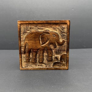 May include: A square, wooden block with a carved relief of an elephant. The elephant is depicted in a detailed, three-dimensional style, with visible tusks and wrinkles. The wood has a warm, brown tone, and the carving is set within a textured frame.