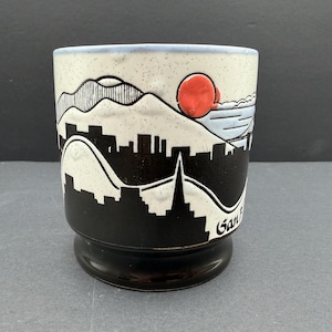 Könnte beinhalten: Keramikbecher mit einem schwarz-weißen Skyline-Design, einer roten Sonne und einer Brücke. Der Becher hat einen gesprenkelten cremefarbenen Hintergrund und einen schwarzen Sockel. Das Wort "San" ist auf der Seite sichtbar.