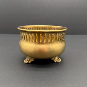 May include: A small, antique brass bowl with a rounded body and three ornate feet. The rim features a series of vertical cutouts. The bowl has a warm, golden hue and is likely used for decorative purposes.