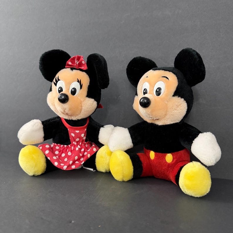 May include: Two plush Disney characters, Mickey and Minnie Mouse, sit side-by-side. Mickey wears red shorts with yellow buttons, while Minnie has a red polka-dot dress. Both have black ears, white gloves, and yellow feet.