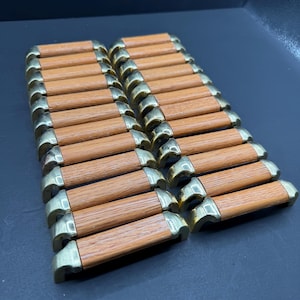 May include: A collection of twenty-two cabinet pulls arranged in two parallel rows. Each pull features a cylindrical light brown wood grain centre section flanked by polished brass metal end caps with curved mounting brackets on each side.