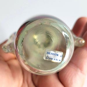 May include: A clear, iridescent glass figurine with a rounded body and two small, curved protrusions. The bottom of the figurine has a sticker that reads "SILVESTR" and "MADE IN TAIWAN". The glass reflects colors of red, purple, and green.