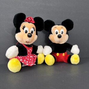May include: Two plush Disney character dolls, Mickey and Minnie Mouse. Minnie wears a red dress with white polka dots and a matching bow. Mickey wears red shorts and yellow shoes. Both have black ears and smiling faces.