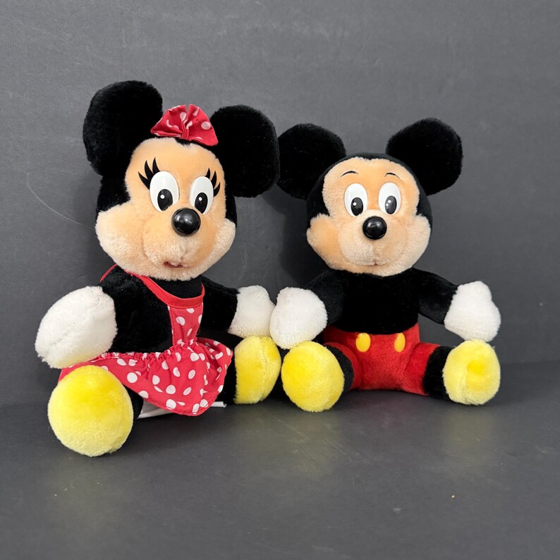 May include: Two plush Disney character dolls, Mickey and Minnie Mouse. Minnie wears a red dress with white polka dots and a matching bow. Mickey wears red shorts and yellow shoes. Both have black ears and smiling faces.