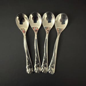 Puede incluir: Cuatro cucharas plateadas con una superficie reflectante dispuestas sobre un fondo negro. Las cucharas tienen un diseño decorativo en la base del mango con estrellas y una figura estilizada. Las cucharas son probablemente para servir o comer.