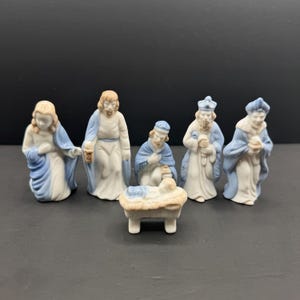 以下が含まれることがあります： 白いローブに青いアクセントが施された、6体の磁器製キリスト降誕フィギュアセット。立ち姿や跪く姿の5体の人物像と、飼い葉桶の中に横たわる小さな赤ちゃんのフィギュアで構成されており、全体に滑らかな釉薬仕上げが施されています。