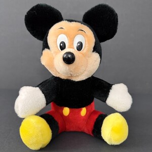 May include: A plush Mickey Mouse toy with a black body, large black ears, and a tan face. The toy has white gloves, red shorts with yellow buttons, and yellow feet. The toy is sitting with its arms and legs spread.