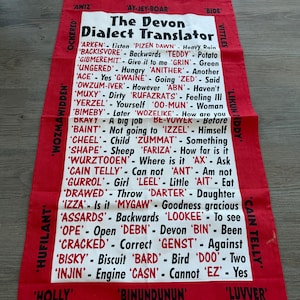 Puede incluir: Paño de cocina rojo y blanco con "The Devon Dialect Translator", una lista de palabras del dialecto de Devon y sus significados. El paño tiene un borde rojo y texto blanco. El texto incluye frases como "Heavy Rain" y "How are you".