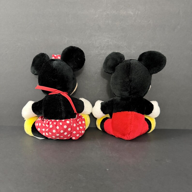 May include: Two plush toys of cartoon characters, viewed from the back. One has a red bow and a red and white polka dot skirt. The other has red shorts. Both have black ears, black bodies, and yellow and white accents.
