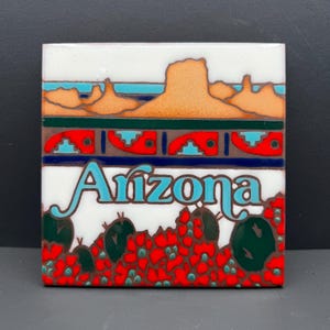 Può includere: Una piastrella quadrata in ceramica con un'illustrazione grafica di un paesaggio dell'Arizona. Il design include mesa arancioni, un cielo turchese, un bordo rosso decorato e un primo piano di cactus verdi con fiori rossi.