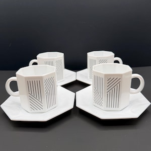 May include: Four white octagonal ceramic cups and matching saucers. Each cup features a geometric pattern of thin black vertical and diagonal lines. The set is arranged in a square formation, showcasing the consistent angular design of the drinkware.