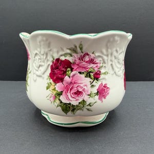 May include: A white ceramic planter with a scalloped rim and base, featuring a green painted edge. The exterior is decorated with embossed scrollwork patterns and a central floral motif of deep red and light pink roses with green leaves.
