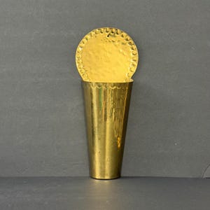 May include: A wall-mounted brass pocket with a polished finish, featuring a semi-circular backplate and a tapered, cone-shaped vessel. The edges of both the circular backplate and the top of the vessel are decorated with a scalloped, hammered pattern.