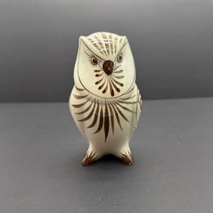 May include: A ceramic owl figurine with a cream body and brown decorative markings. The owl has large eyes and a detailed feather pattern. The figurine is standing upright and has a small, rounded base.