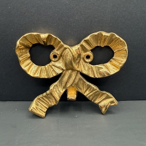 May include: A gold-toned metal wall hook shaped like a decorative ribbon bow. The bow features a textured, pleated surface with two circular screw holes positioned in the centre for mounting. A curved hook extends downward from the centre knot.