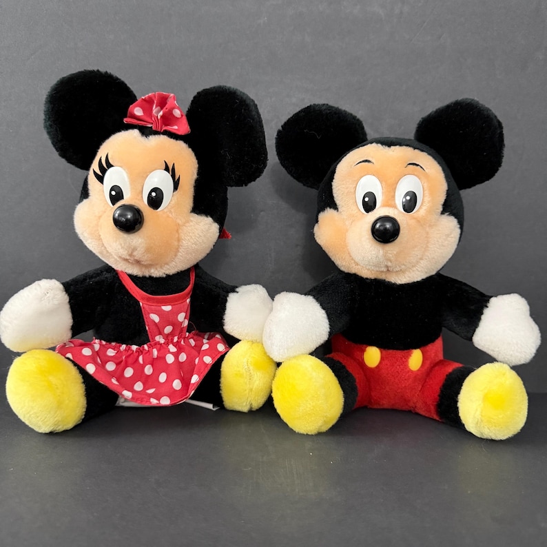 May include: Two plush Disney characters, Mickey and Minnie Mouse, sit side-by-side. Mickey wears red shorts and yellow shoes. Minnie wears a red polka-dot dress, a red bow, and yellow shoes. Both have black ears, black fur, and tan faces.