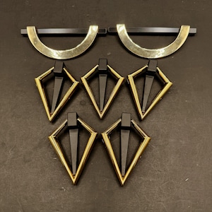 May include: A collection of furniture hardware consisting of two semi-circular gold and black drawer pulls and five diamond-shaped gold and black drop pulls. Each piece features a metallic gold frame with a solid black centre component.